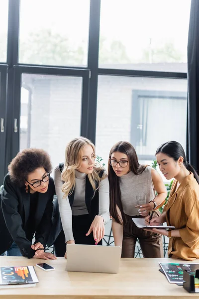 Blonde businesswoman pointing at laptop near young multiethnic colleagues in office — Stock Photo