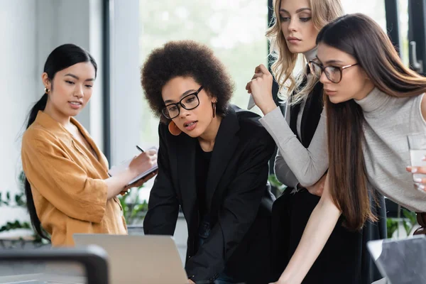 Asian businesswoman writing in notebook near multicultural colleagues looking at laptop — Stock Photo