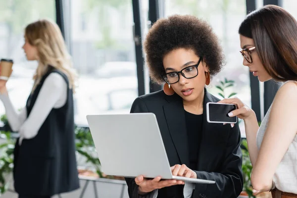 Mujer de negocios afroamericana mirando portátil cerca de colegas con teléfono inteligente en la oficina - foto de stock