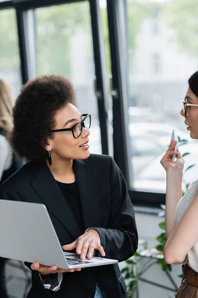 African american businesswoman in eyeglasses using laptop while talking to colleague — Stock Photo