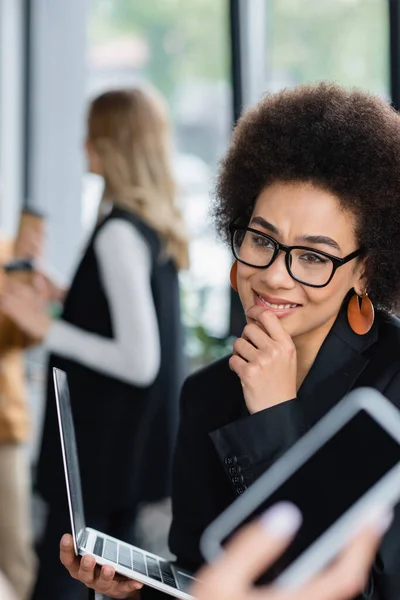 Афроамериканська бізнесменка в окулярах посміхається близьким розмитим колегам зі смартфоном. — стокове фото