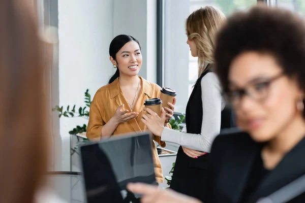 Interracial businesswomen with paper cups talking in office during coffee break — Stock Photo