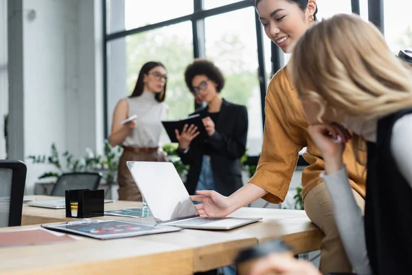 Sonriente asiático mujer de negocios mirando portátil cerca borrosa colega en oficina - foto de stock