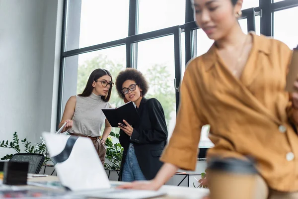 Interracial colleagues in eyeglasses looking at notebook near blurred asian businesswoman — Stock Photo