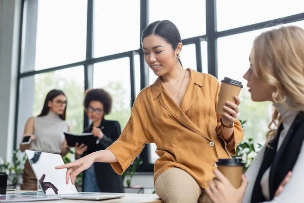 Asian manager pointing at laptop while sitting on desk with paper cup near blurred colleague — Stock Photo