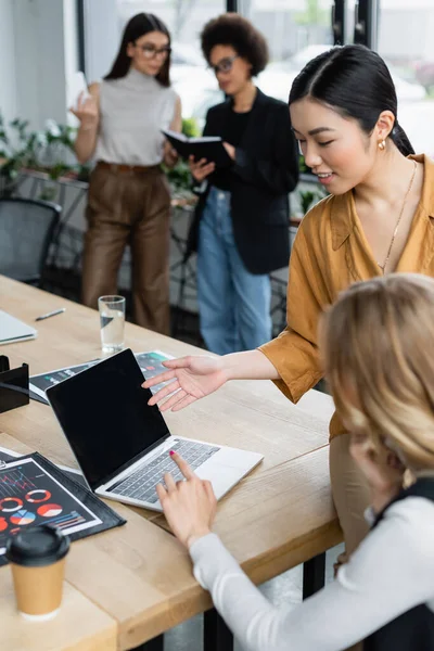 Asiático mujer de negocios apuntando a portátil cerca borrosa manager y infografías - foto de stock