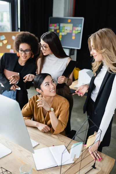 Happy multiethnic businesswomen pointing at computer monitor while working in office — Stock Photo