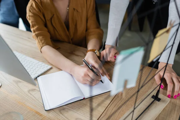 Partial view of businesswoman writing in empty notebook near colleague at workplace — Stock Photo