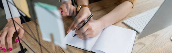 Cropped view of businesswoman writing in blank notebook near colleague, banner — Stock Photo