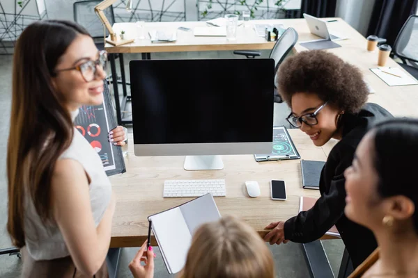 Blurred businesswoman holding empty notebook near smiling colleagues and monitor with blank screen — Stock Photo