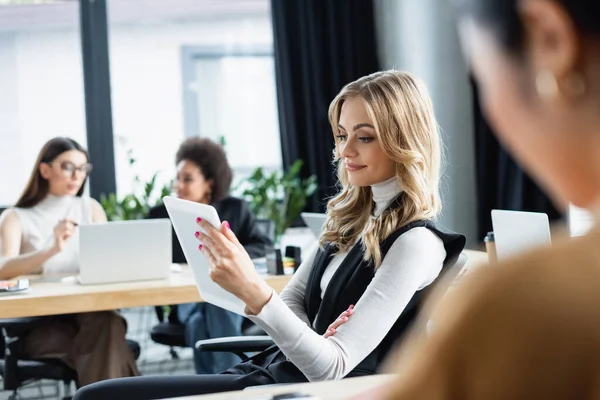 Blonde Managerin lächelt, während sie digitales Tablet in der Nähe verschwommener multiethnischer Mitarbeiter benutzt — Stockfoto