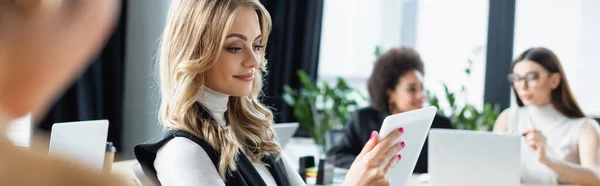 Улыбающаяся деловая женщина, глядя на цифровой планшет вблизи размытых мультиэтнических коллег в офисе, баннер — стоковое фото
