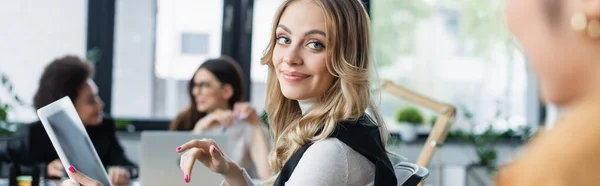 Sonriente mujer de negocios apuntando a la tableta digital con pantalla en blanco cerca de colegas borrosos, pancarta - foto de stock