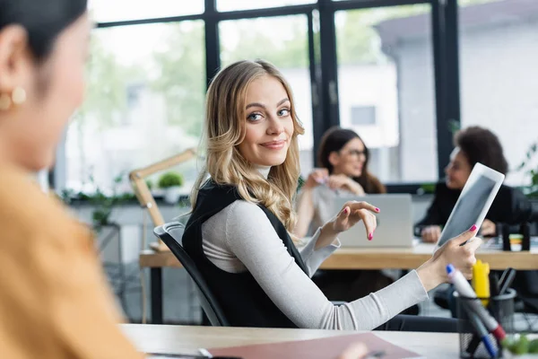 Blonde businesswoman with digital tablet smiling at colleague on blurred foreground — Stock Photo