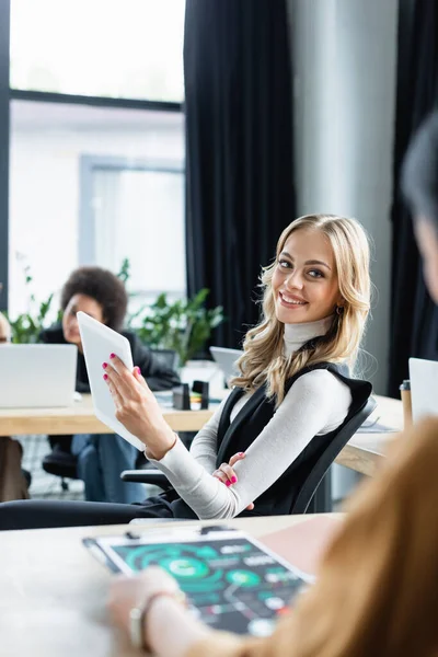 Blonde businesswoman with digital tablet smiling near colleague on blurred foreground — Stock Photo