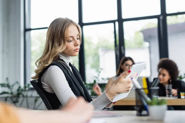 Manager bionda guardando tablet digitale vicino a colleghi interrazziali che lavorano su sfondo sfocato — Foto stock