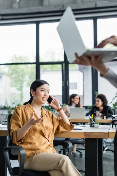 Smiling asian woman gesturing while talking on mobile phone near blurred multiethnic colleagues — Stock Photo