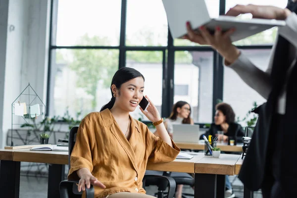 Asian businesswoman talking on mobile phone near blurred colleague with laptop — Stock Photo