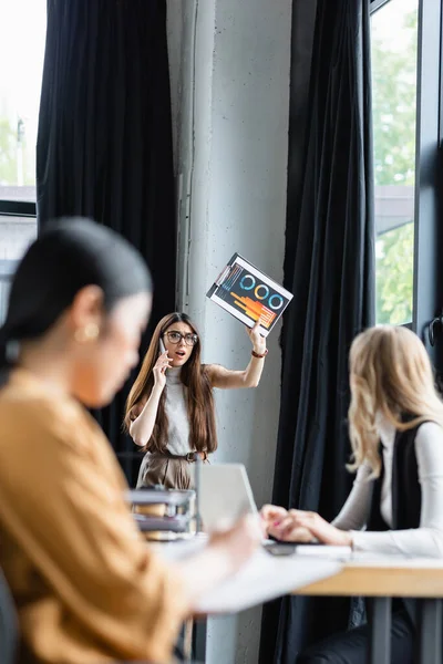 Excitada empresária acenando com infográficos enquanto falava no smartphone perto de colegas embaçados — Fotografia de Stock