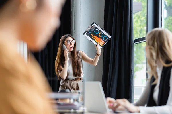 Збуджена бізнес-леді махає буфера з графіками під час розмови на смартфоні поруч з розмитими колегами — стокове фото