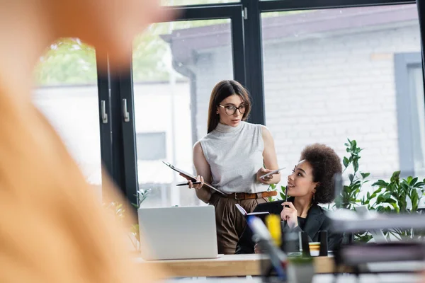 Young interracial businesswomen talking in office on blurred foreground — Stock Photo