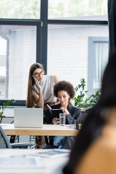 Brunette woman talking on cellphone near african american colleague with notebook in office — Stock Photo