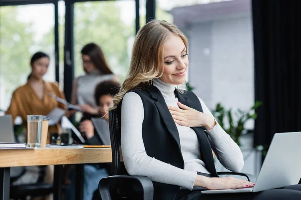 Pleased businesswoman touching chest while working on laptop near blurred multicultural coworkers — Stock Photo