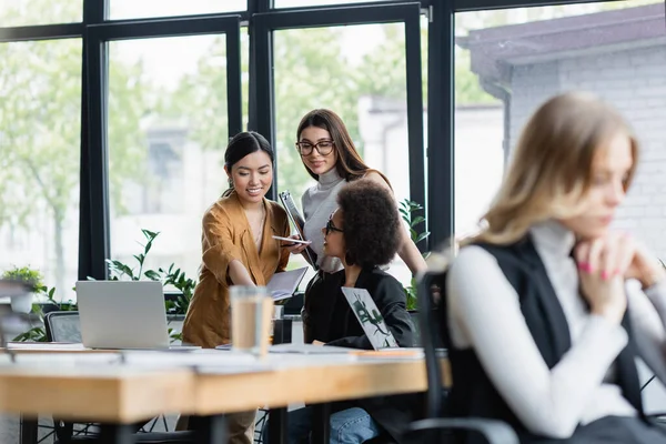 Mujeres de negocios multiétnicas positivas hablando cerca de ventana en la oficina en primer plano borrosa - foto de stock