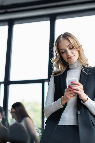 Mensajería rubia de mujer de negocios en el teléfono inteligente cerca de colegas en un fondo borroso - foto de stock