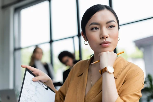 Dreamy asian fashion designer looking away while holding clipboard with drawing — Stock Photo