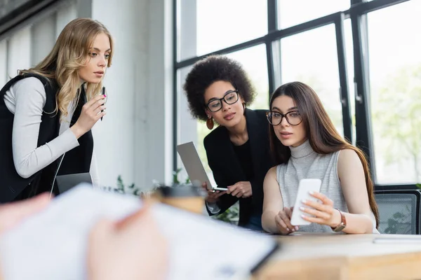 Jeune femme d'affaires pointant vers le smartphone tout en travaillant avec des collègues interraciaux au premier plan flou — Photo de stock