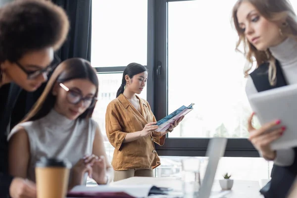 Junge asiatische Geschäftsfrau liest Magazin, während Kollegen an verschwommenem Vordergrund arbeiten — Stockfoto