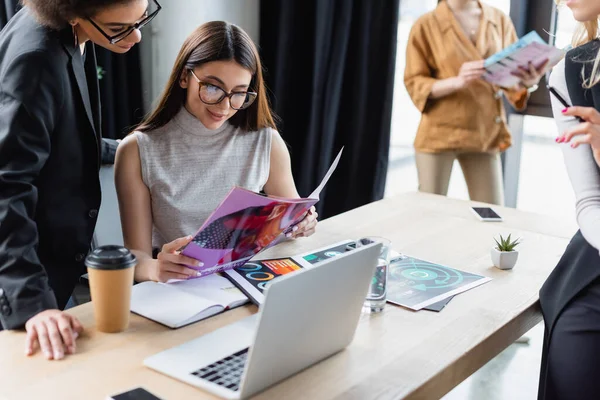 Rivista di lettura business donna positiva vicino laptop e collega afro-americano — Foto stock