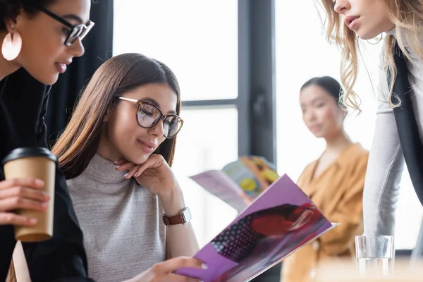 Brunette femme d'affaires souriant tout en lisant le magazine près de collègues multiethniques — Photo de stock