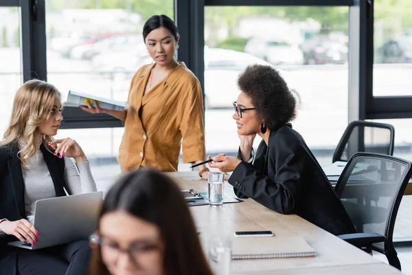 Afrikanisch-amerikanische Geschäftsfrau zeigt mit Stift, während sie mit Kollegin mit Laptop spricht — Stockfoto