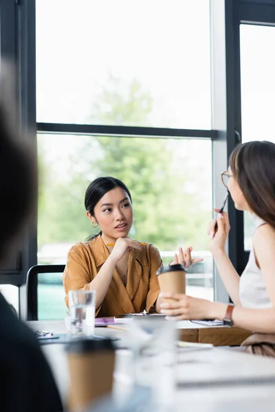 Junge asiatische Geschäftsfrau zeigt mit dem Finger, während sie mit Kollegin auf verschwommenem Vordergrund spricht — Stockfoto