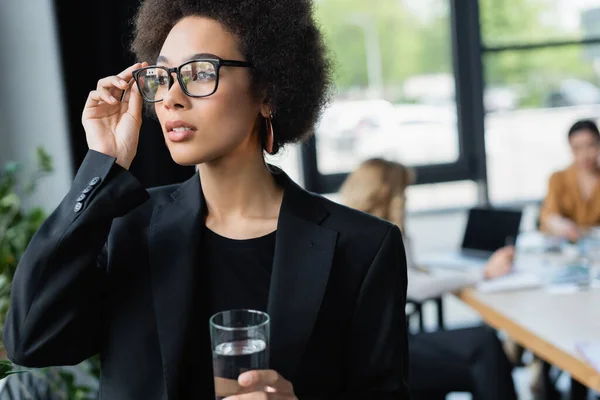 Jeune femme d'affaires afro-américaine touchant des lunettes tout en se tenant debout avec un verre d'eau — Photo de stock