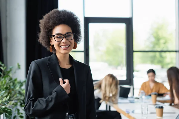 Жизнерадостная африканская американская бизнесвумен улыбается в камеру рядом с размытыми менеджерами в офисе — стоковое фото