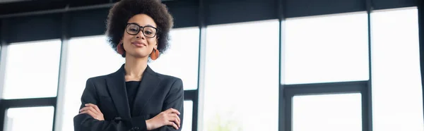 Femme d'affaires afro-américaine en lunettes debout avec les bras croisés au bureau, bannière — Photo de stock