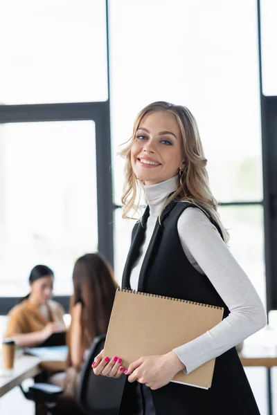Улыбающаяся деловая женщина смотрит в камеру, стоя с бумажной папкой в офисе — стоковое фото