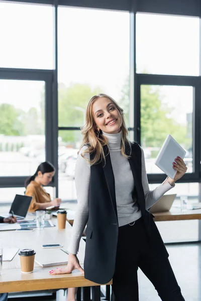 Fröhliche Geschäftsfrau mit digitalem Tablet blickt in die Kamera neben Kollegen, die an verschwommenem Hintergrund arbeiten — Stockfoto