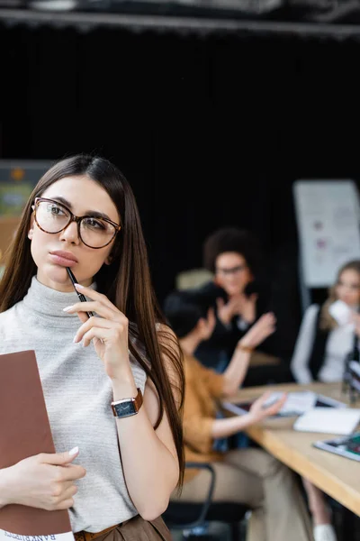 Mujer de negocios con carpeta de papel y lápiz pensando cerca de los gerentes que trabajan sobre fondo borroso - foto de stock