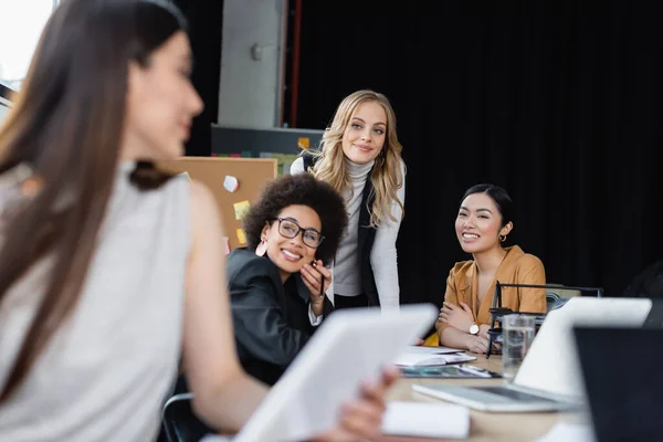 Cheerful multiethnic businesswomen looking at blurred colleague with digital tablet on blurred foreground — Stock Photo