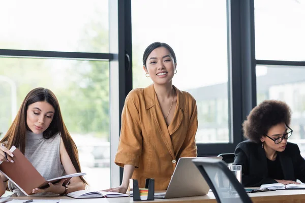 Happy asian manager smiling at camera near interracial colleagues at workplace — Stock Photo