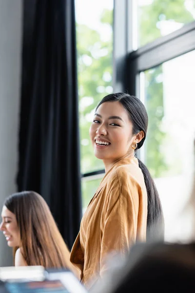 Freudiger asiatischer Manager lächelt in die Kamera neben verschwommenen Kollegen im Büro — Stockfoto