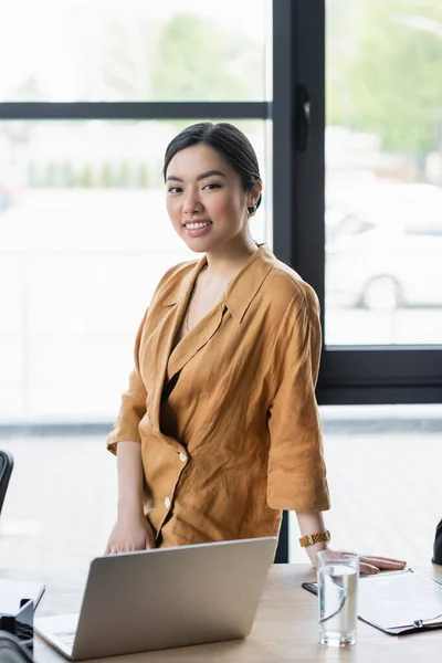 Heureux asiatique femme d'affaires souriant à caméra près ordinateur portable sur le bureau dans le bureau — Photo de stock