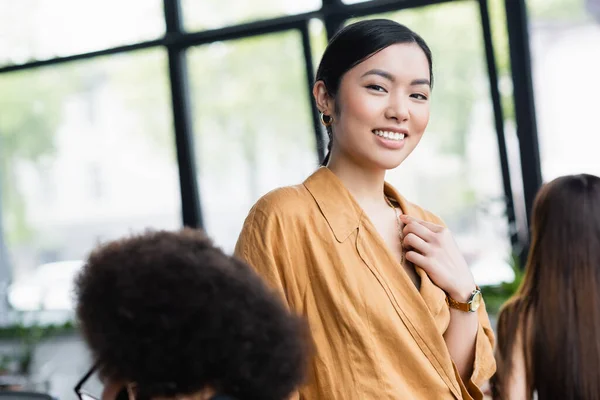 Stylische asiatische Geschäftsfrau lächelt in die Kamera neben verschwommenen Kollegen — Stockfoto