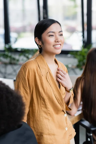 Joyeuse asiatique femme d'affaires regardant loin tout en se tenant près de collègues sur flou premier plan — Photo de stock