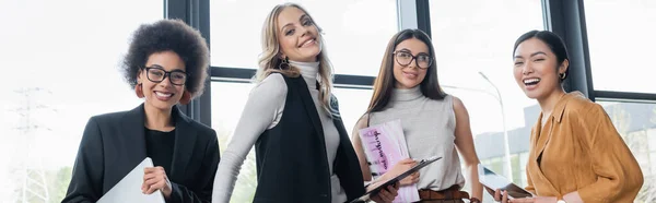 Aufgeregte multiethnische Manager mit Gadgets und Dokumenten, die in die Kamera im Büro schauen, Banner — Stockfoto