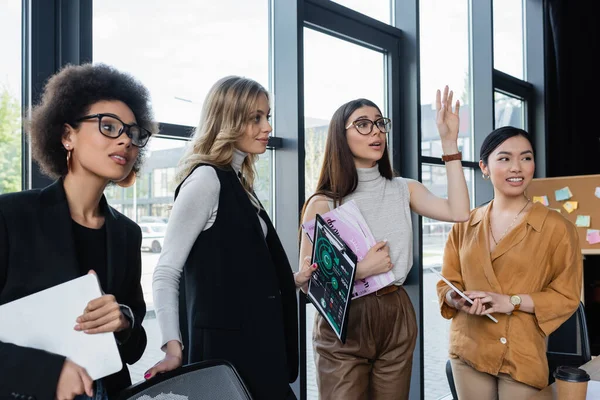 Mujer de negocios en gafas de vista saludando de la mano mientras mira hacia otro lado cerca de socios comerciales multiétnicos en la oficina - foto de stock